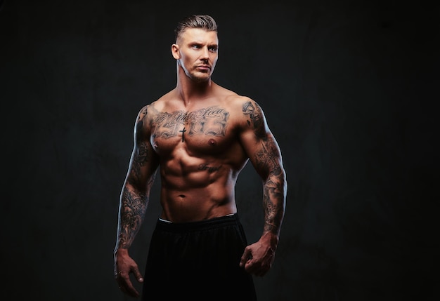 A tattooed muscular shirtless man with stylish hair posing at the camera on a dark background.