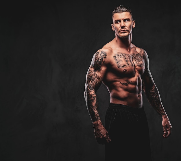 A tattooed muscular shirtless man with stylish hair posing at\
the camera on a dark background.