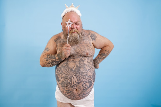 Photo tattooed obese man with crown looks through star on magic stick on blue background