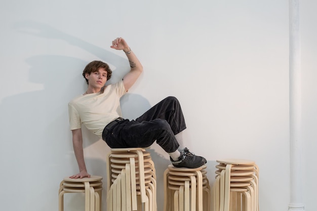 Tattooed Young man sitting on the top of rack of stools and leaning on the white wall