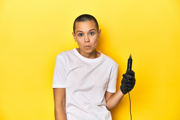 Tattooist woman in studio shaved head yellow background shrugs shoulders and open eyes confused