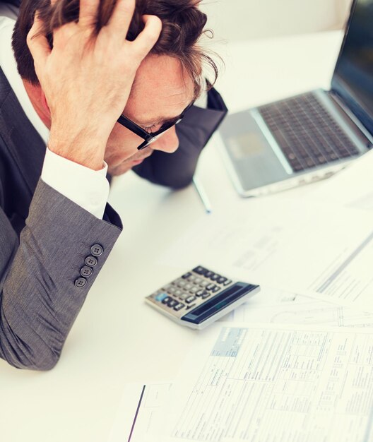 tax, business and stress concept - stressed businessman with papres, laptop and calculator