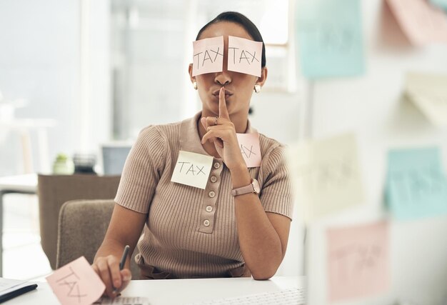 Photo tax evasion is a crime shot of a young businesswoman covered in sticky notes and holding her fingers on her lips while working on taxes in an office