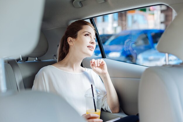 Taxi ride. Thoughtful smart attractive businesswoman sitting in the car and holding a cup with juice while going for work