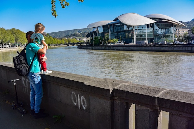 Tbilisi georgia april 27 2019 a young man with a child on the embankment of the kura river mtkvari with the tbilisi public service hall located on the bank in the heart of the city