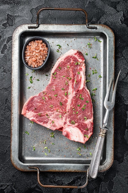 Tbone steak raw marbled beef meat in a steel kitchen tray with herbs Black background Top view