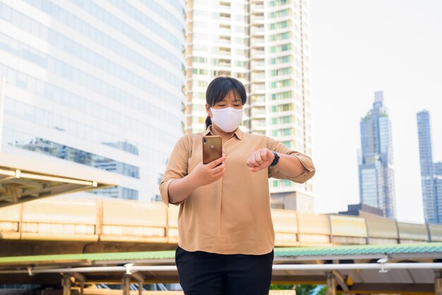 Te zware Aziatische vrouw met masker gebruikend telefoon en controlerend de tijd bij skywalkbrug