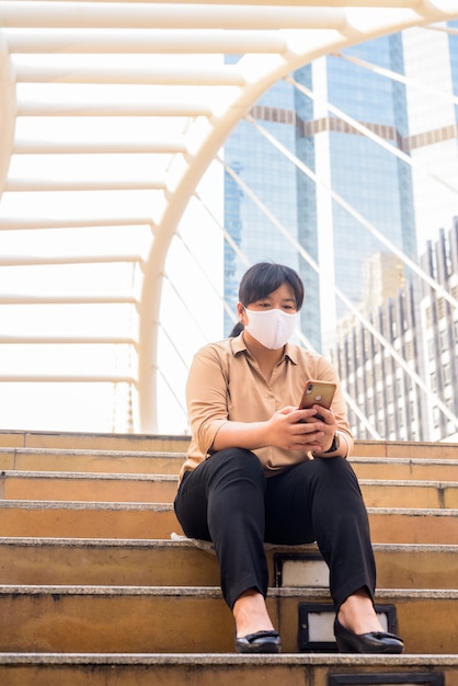 Te zware Aziatische vrouw met masker gebruikend telefoon en zittend in de stad