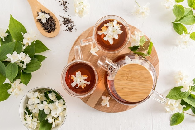 Composizione del tè su sfondo bianco una teiera di vetro e due tazze di vetro di tè al gelsomino su una tavola rotonda di legno tra fiori di gelsomino