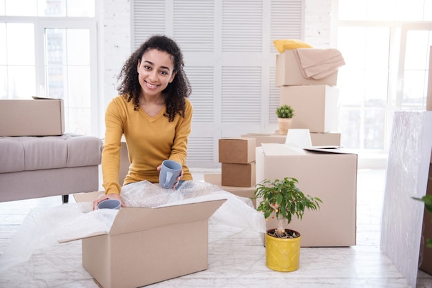 Tea drinker. Charming young woman putting tea cups into the box and smiling at the camera while packing her belongings before moving out