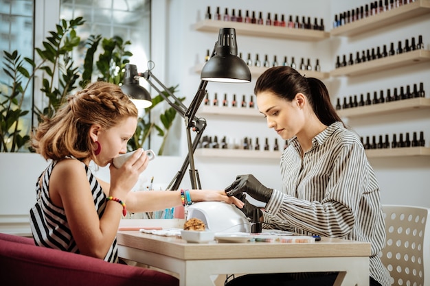 Tea during manicure. Stylish teenage girl wearing accessories and striped dress drinking tea during manicure