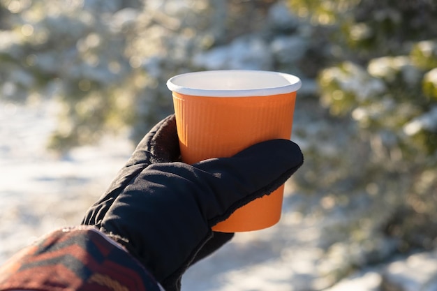찻잔 겨울 여행. 여성의 장갑을 낀 손에 있는 주황색 종이컵. 겨울 밝고 맑은 배경