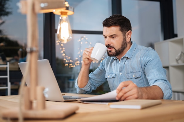 Tea time. Nice pleasant hard working businessman looking at the laptop screen and having tea while doing his job