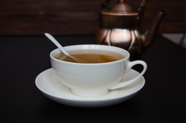 Tea in white cup with saucer and tea pot on a dark background