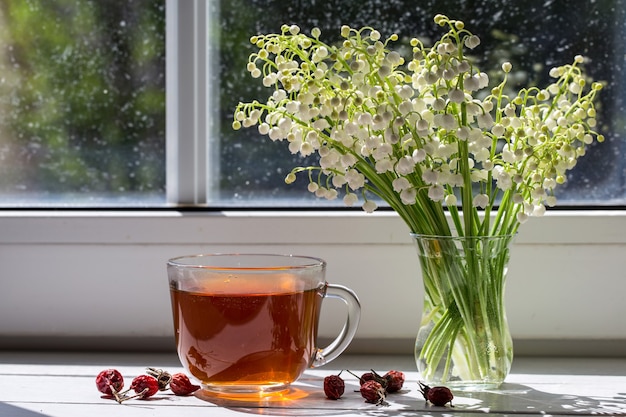 Tea with a dogrose and a bouquet of spring lilies of the valley on the windowsill.
