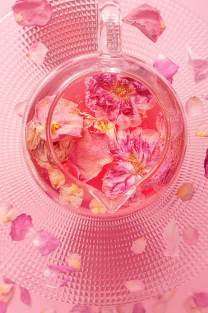 Tea with rose petals in glass teapot on pink background