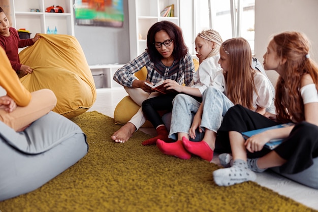 Teacher in glasses sitting on a pouf and reading for children in class