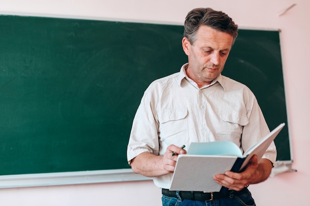 Teacher holding and reading textbook standing next a blackboard.