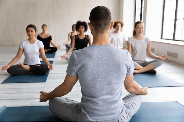 Teacher instructor training diverse young people rear view doing Padmasana exercise practicing yoga at group lesson sitting in Lotus pose on mat working out meditating in modern fitness center