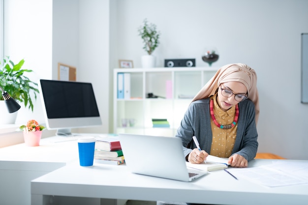 Teacher near laptop. Young Muslim teacher wearing glasses sitting near laptop and writing notes