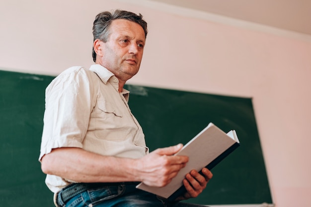teacher sitting with open textbook next a blackboard.