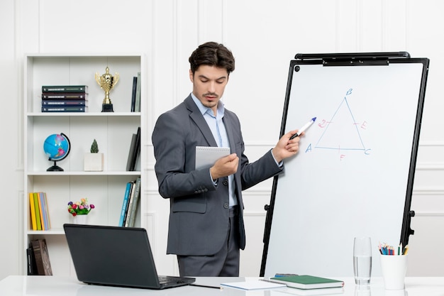 Teacher smart instructor in grey suit in classroom with computer and whiteboard looking at notes