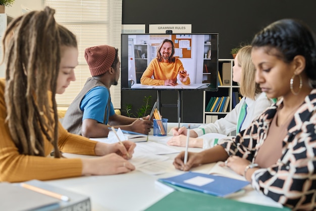 L'insegnante insegna ai suoi studenti online durante la chiamata video sullo schermo mentre sono seduti alla scrivania e fanno appunti nel quaderno