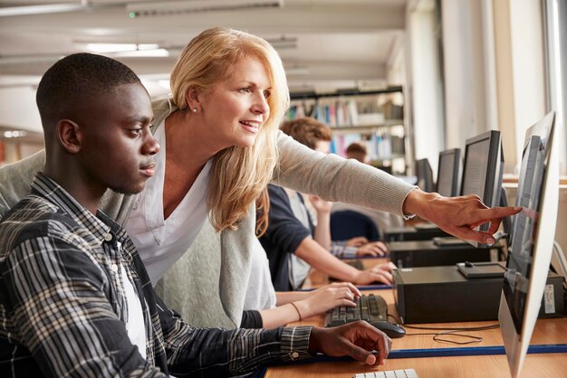 Teacher With Male Student Working On Computer In College Library