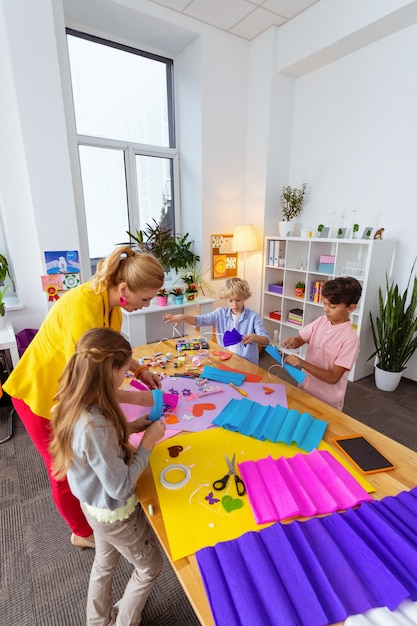 Teacher with pupils. Blonde-haired teacher standing near pupils making cutouts with colorful paper