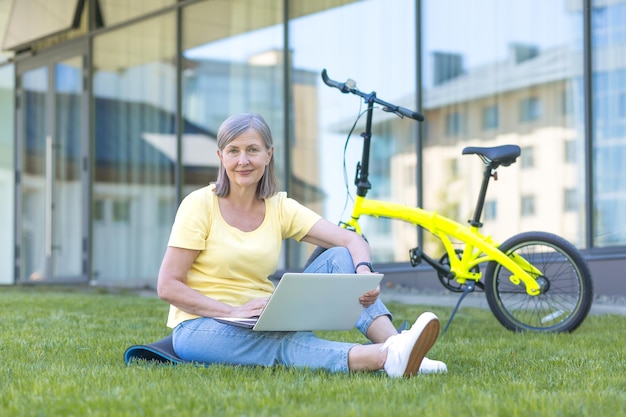 노트북과 자전거로 풀밭에 앉아 있는 아름다운 고위 여성의 초상 그는 공부하고 일하며 카메라 미소를 바라보고 있다