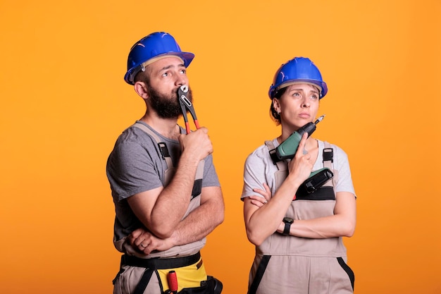 Team of builders brainstorming construction ideas in studio, thinking about renovating solution to start rebuilding work. Pensive man and woman holding power drilling gun and pair of pliers.