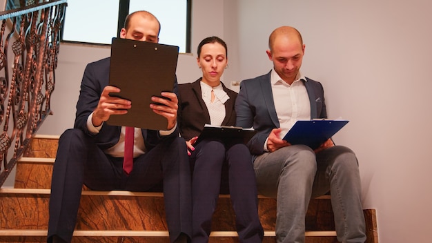 Team of business people making plans analysing annual graphs enjoying sitting on staircase explaining finance project. Group of company manager and office executive on busy stairs of workplace.