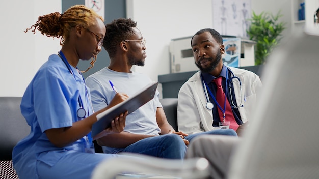 Team of clinical specialists talking to patient at hospital reception, doing medical consultation with young man. Physician and nurse explaining disease and medicare to person. Handheld shot.