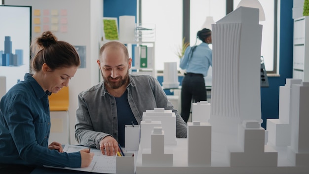 Team of coworkers analyzing building model and blueprints plan\
on table to design construction layout. architectural colleagues\
doing teamwork, designing project development together.