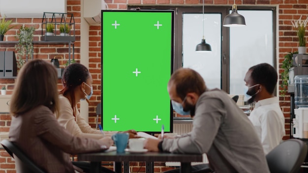 Team of diverse workmates working with vertically green screen on monitor, wearing face mask. Business people using isolated chroma key with copy space and blank mock up background.