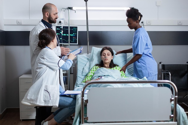 Team of doctors discussing patient medical history on clipboard\
before surgery while nurse is giving medical care. woman with low\
spo2 saturation connected to iv drip line and monitor showing\
vitals.