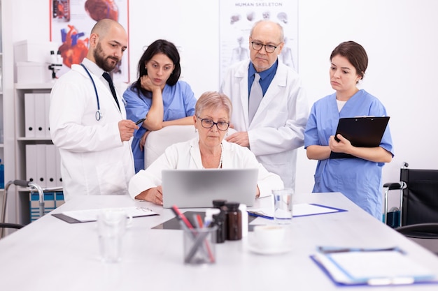 의료 유니폼을 입고 회의실에서 노트북을 보고 있는 의사 팀. 질병에 대해 동료와 이야기하는 클리닉 전문 치료사, 의학 전문가