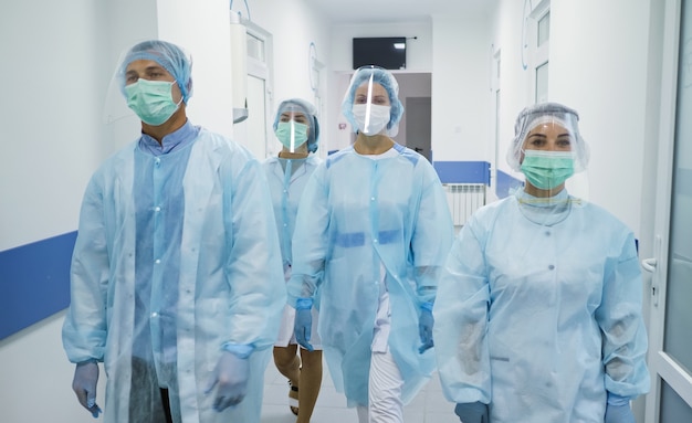 Бригада врачей в защитных костюмах медицинских работников в масках идет по коридору современного госпиталя ...