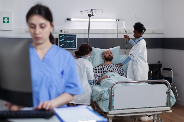 Team of doctors setting up iv drip line and consulting patient connected to monitor measuring vitals while nurse reads lab results. Health care worker writing admission while doctors examine ill man.