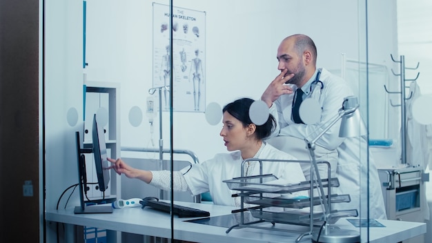 Team of doctors talking over a glass wall, pointing to PC screen while people are walking in the hallway. Healthcare system, private modern medical hospital clinic