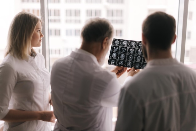 Team of experts doctors discusses the results of an MRI scan of the patient's head in consulting room of hospital against large window. View from the doctors' backs. Concept of team medical work.