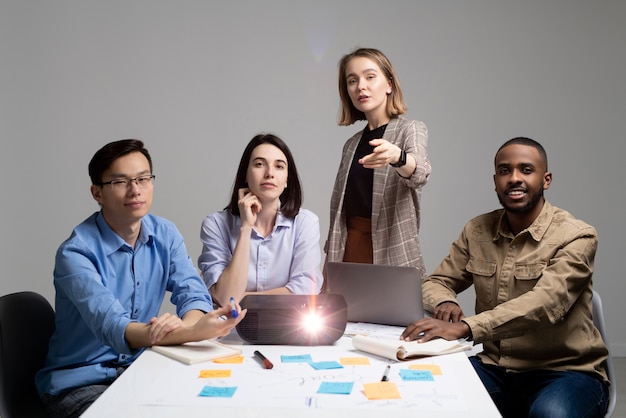 Team leader pointing at camera while inviting you to become part of startup team, group of specialists sitting at table with projector