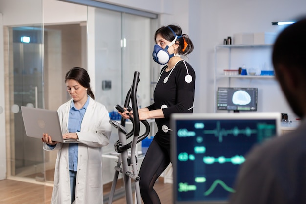 Team of medial researchers monitoring vo2 of woman performance sports wearing mask running. Lab science doctor measuring endurance using tablet while ekg scan runs on computer screen in laboratory.