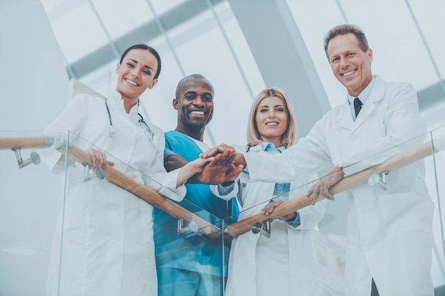 Photo team of medical experts. low angle view of four happy medical doctors standing close to each other and holding their hands together while leaning at the handrail