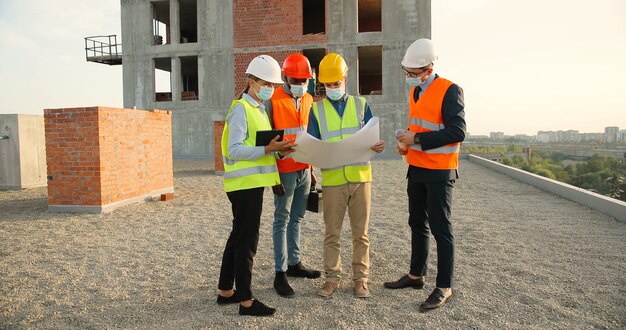 Squadra di costruttori di razze miste maschi e femmine con elmetti e maschere mediche in piedi in cima all'edificio e discutendo della bozza del piano di costruzione. concetto di pandemia. ingegneri e architetti.