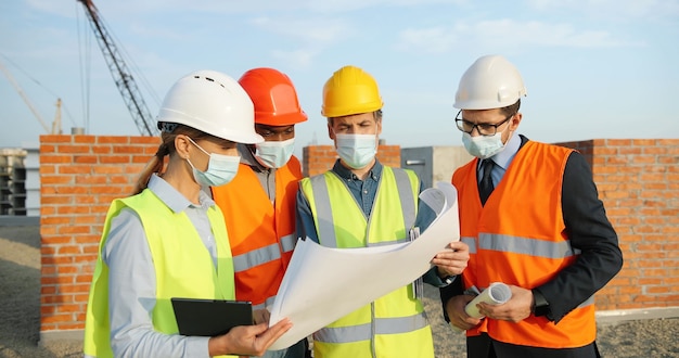 建物の最上部に立って計画案の作成について話し合っている、ヘルメットと医療用マスクの混血の男性と女性の建設業者のチーム。コロナウイルスの概念。エンジニアと建築家。