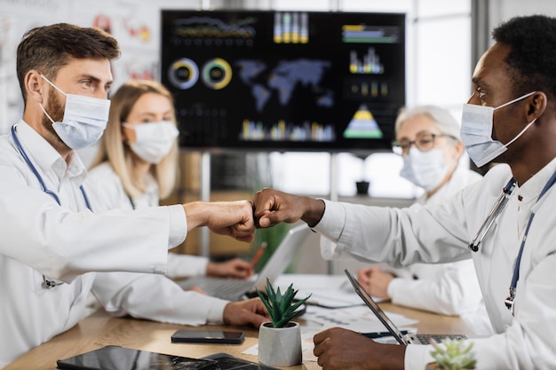 Photo team of physicians in face masks giving fist bump indoor