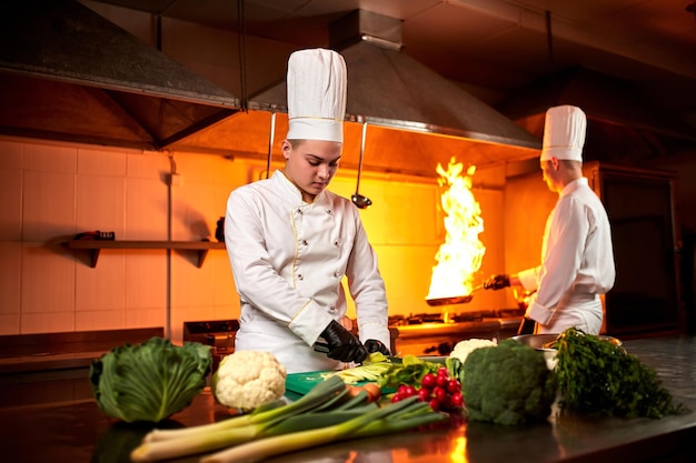 A team of professional chefs cook meals with frying pan and\
fire in the kitchen of restaurant chief chef preparing dish using\
different food ingredients vegetables meat and fish