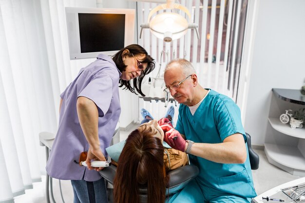 Team of professional male doctor dentist and his female assistant doing examination and treatment to young female patient. Healthcare, dentistry concept.