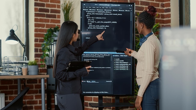 Team of programmers analyzing code on wall screen tv looking\
for bugs and errors while holding digital tablet and remote\
control. software engineers collaborating on cloud computing group\
project.